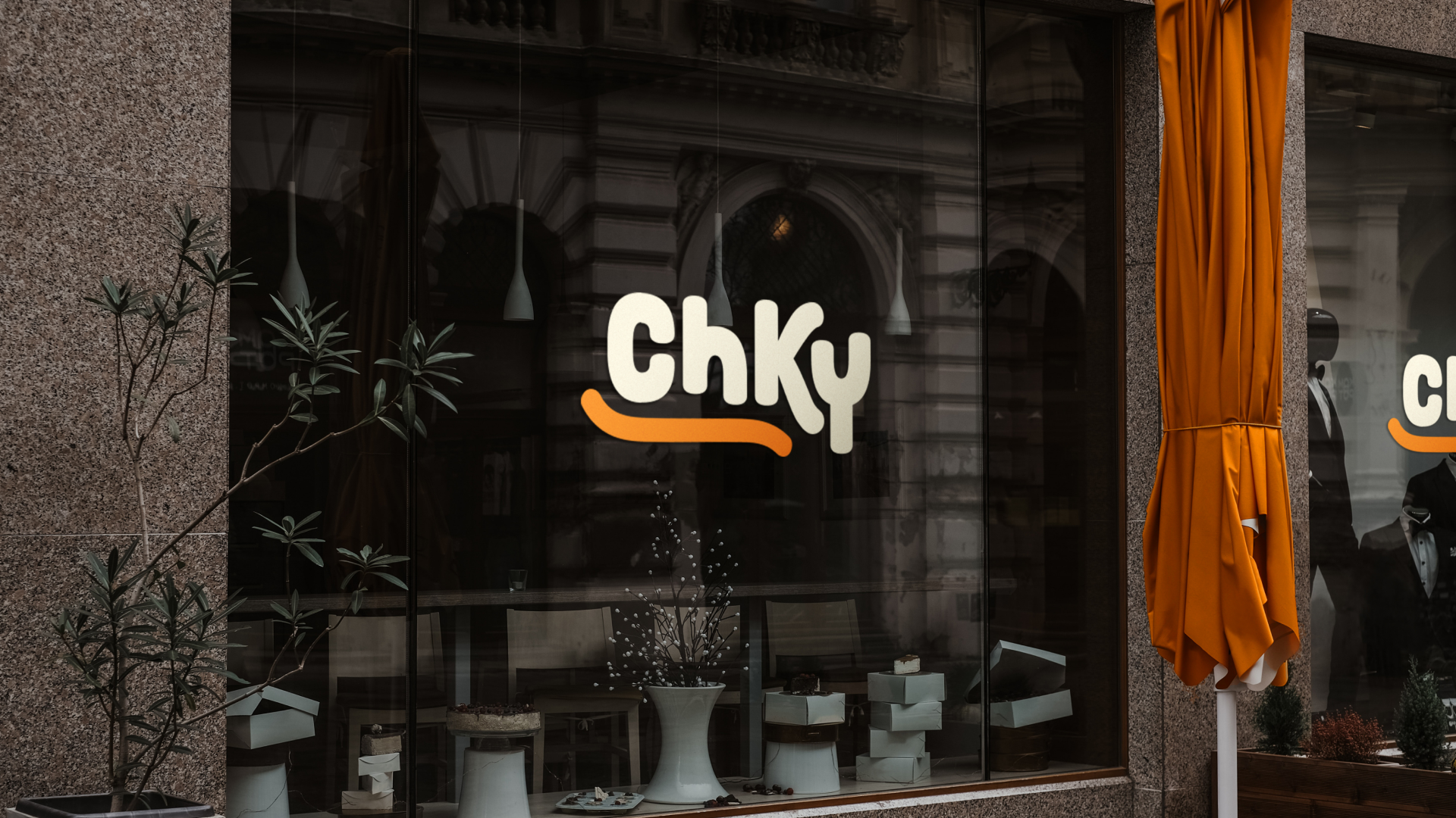Storefront with a large window displaying the ChKY logo in bold letters. Inside, various items are showcased on white display stands, reflecting a design subscription flair. An orange umbrella is folded near the entrance of the stone building.