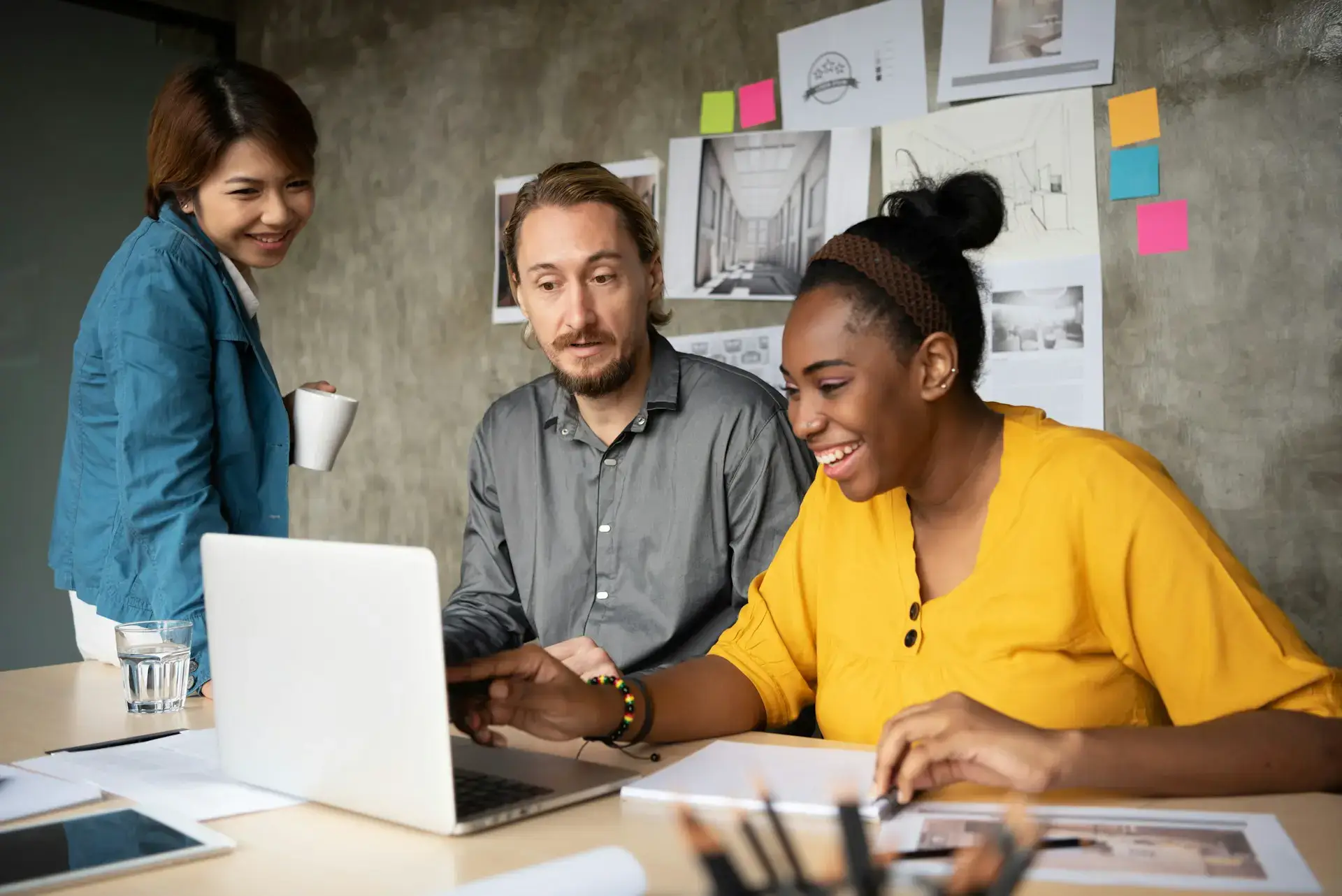 Three colleagues in a creative office setting gather around a laptop. A woman in a yellow shirt smiles, pointing at the screen showcasing their unlimited graphic design project. A man in a gray shirt looks on, while a woman in a teal jacket stands with a cup, smiling. Plans are on the wall.