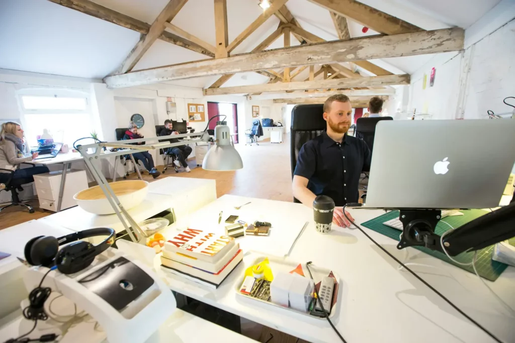 A spacious, bright office with wooden beams and several people working at desks. A man in the foreground is using a laptop, likely managing affiliate programs. Various office supplies are scattered around, and natural light fills the room through windows.
