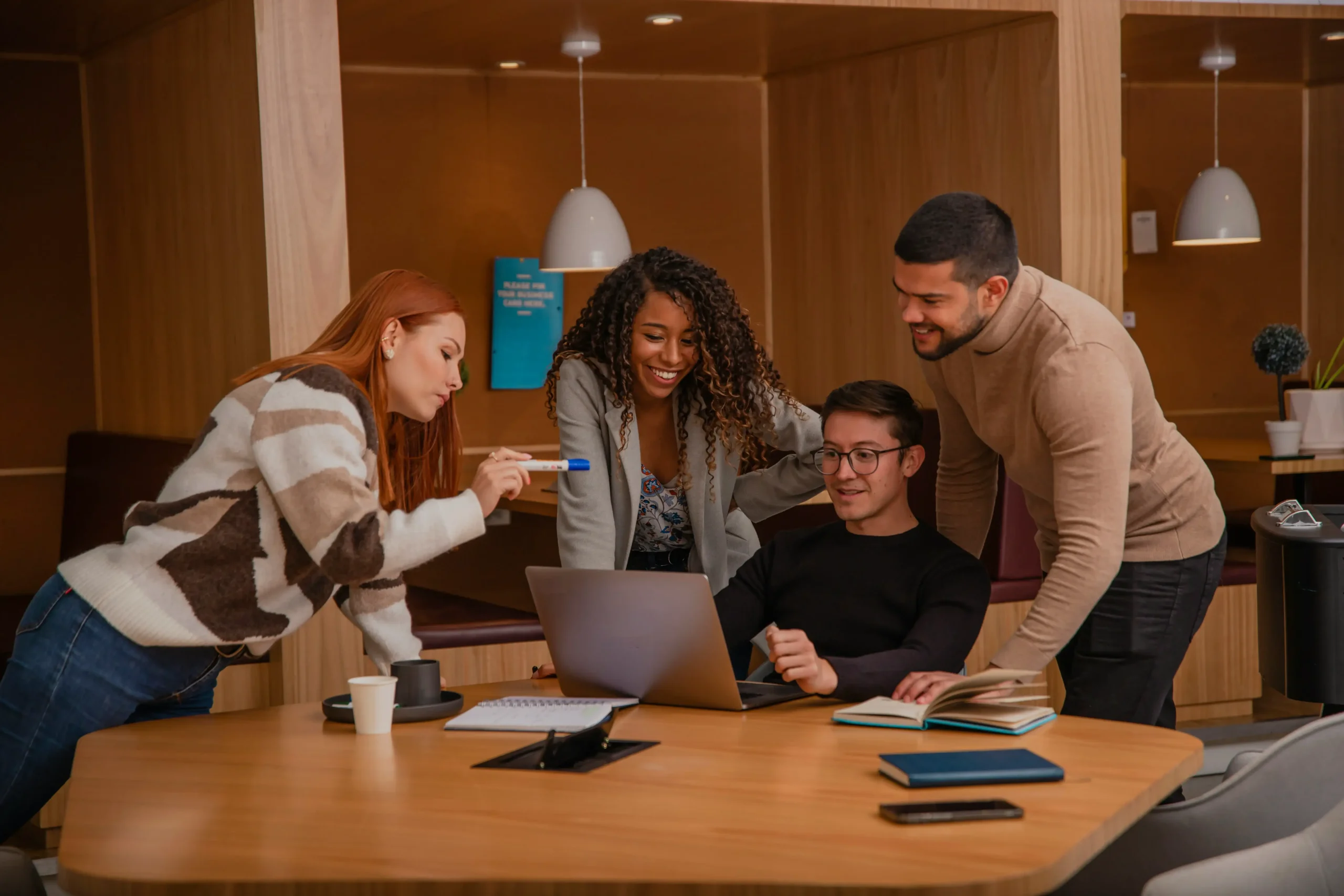 A group of four people in a cozy office setting gather around a laptop, engaged and smiling. One person sits at the table, while the others stand around, with books and a coffee cup also visible on the table.