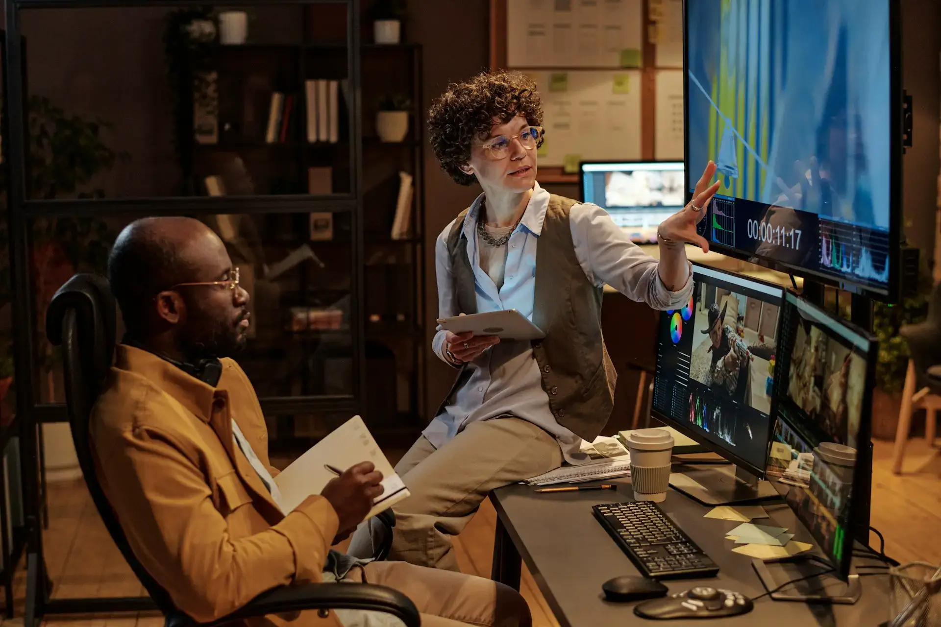 Two people in an office work on video editing. One points at a monitor displaying video editing software, while the other takes notes. A tablet, keyboard, and coffee cup are on the desk. The room is dimly lit, creating a focused atmosphere.