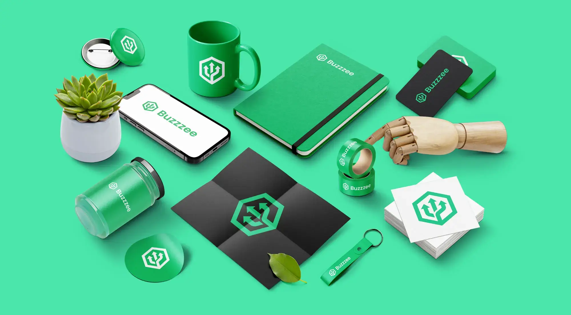 A flat lay of various branded items on a green surface, including a mug, notebook, smartphone, coaster, jar, business cards, and a small potted plant. A wooden mannequin hand holds a green ball, all featuring the BUZZEO logo.