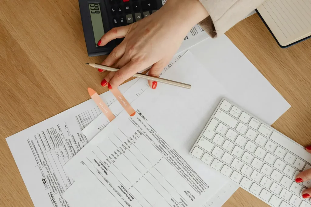 A person is using a calculator while holding a pencil over various financial documents spread on a wooden desk. A computer keyboard is partially visible on the right. The person has red nail polish, and some sticky notes are nearby.